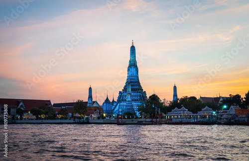 Beautiful stupa of Wat Arun decorated in blue lights at sunset seen from a ferry boat on Chao Phraya river, Bangkok, Thailand