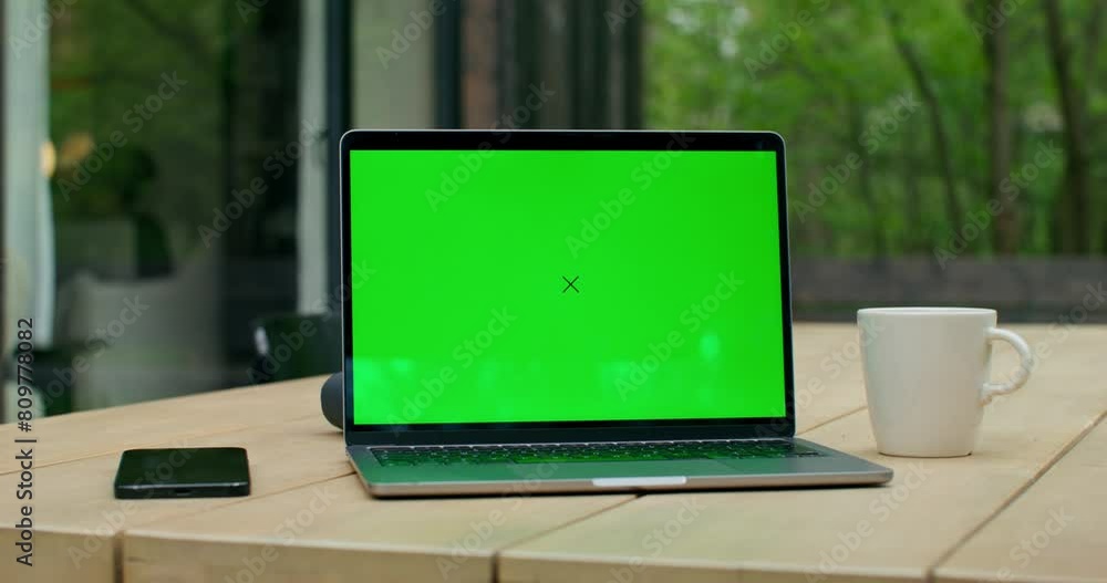 An open laptop with a green screen is on a table on the terrace of a stylish cottage. An open laptop with a green screen is on a table on the terrace of a stylish cottage.