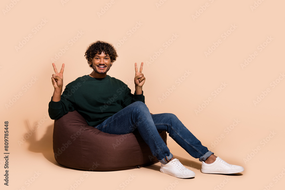 Full length photo of cheerful guy dressed sweatshirt sitting bean bag showing v-signs emtpy space isolated beige color background Full length photo of cheerful guy dressed sweatshirt sitting bean bag showing v-signs emtpy space isolated beige color background