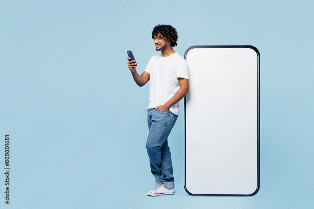 © ViDi Studio - Full body side view young fun Indian man wear white t-shirt casual clothes big huge blank screen mobile cell phone with area use smartphone isolated on plain pastel blue background. Lifestyle concept. © ViDi Studio - Full body side view young fun Indian man wear white t-shirt casual clothes big huge blank screen mobile cell phone with area use smartphone isolated on plain pastel blue background. Lifestyle concept.