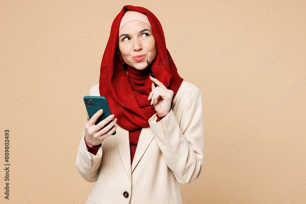 Young minded Arabian Asian Muslim woman wear red abaya hijab suit clothes hold use mobile cell phone look aside isolated on plain beige background studio. UAE middle eastern Islam religious concept. Young minded Arabian Asian Muslim woman wear red abaya hijab suit clothes hold use mobile cell phone look aside isolated on plain beige background studio. UAE middle eastern Islam religious concept.