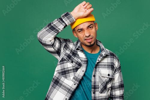 Young sad ill tired exhausted man of African American ethnicity he wears shirt blue t-shirt yellow hat look camera spread hands isolated on plain green background studio portrait. Lifestyle concept.