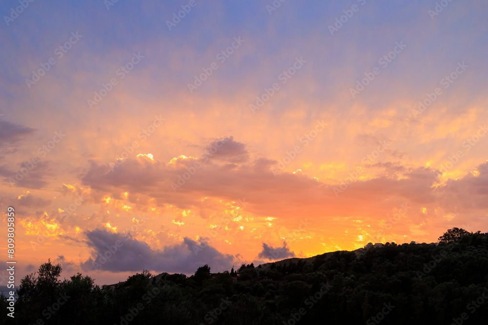 Fototapeta premium Colourful sunset in the bay of Agios Georgios with dark clouds in front of an intensely orange sky with individual strangely shaped dark wispy clouds on the island of Corfu
