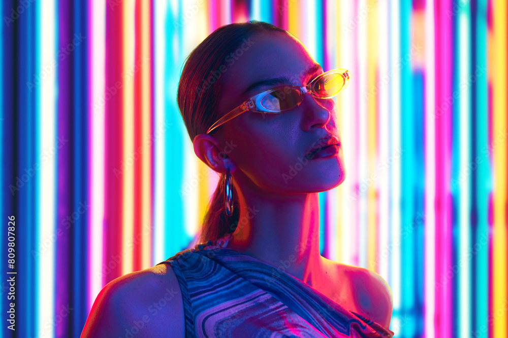 Modern, stylish woman in sunglasses, set against backdrop illuminated by striking neon lights in shades of red and pink. Futuristic look. Concept of youth, beauty, fashion and style, modern lifestyle.