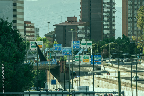 Urban entrance road to Madrid with several lanes in each direction and a multitude of large signs indicating directions and exits from the highway between buildings and urban residences