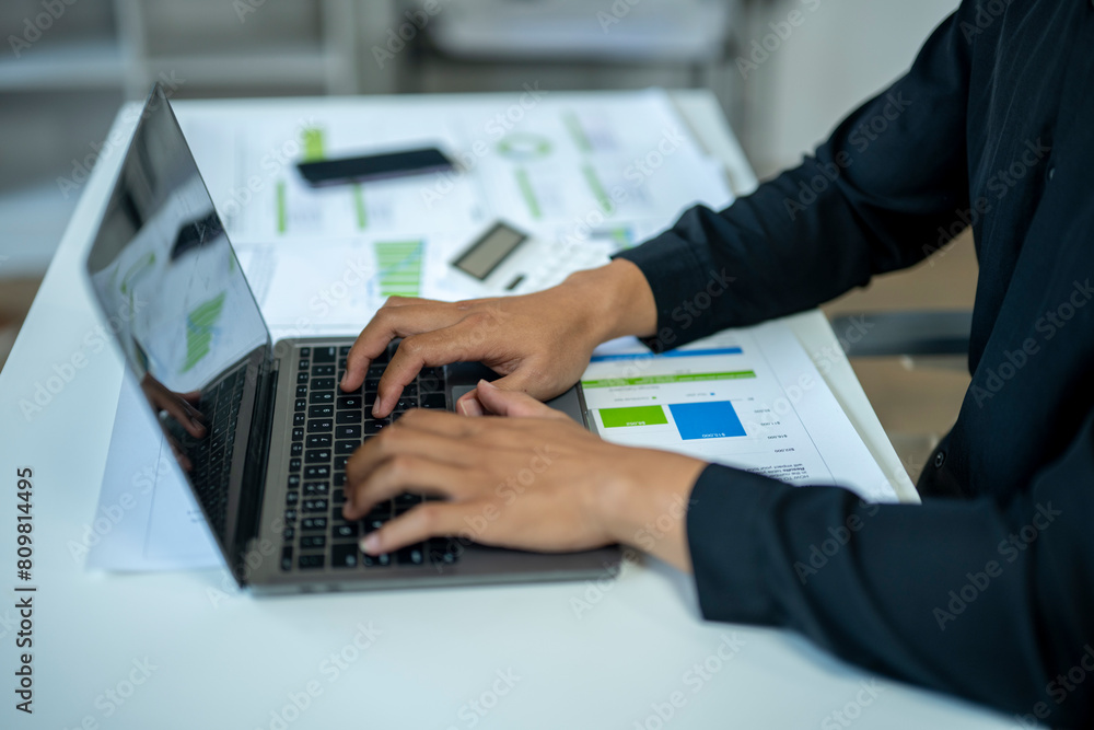 A person is typing on a laptop with a green and blue graph on the screen. The person is wearing a black suit and he is working on a project A person is typing on a laptop with a green and blue graph on the screen. The person is wearing a black suit and he is working on a project