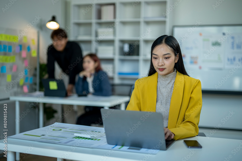 A woman in a yellow jacket is working on a laptop in front of a white board. She is surrounded by other people in the room, some of whom are looking at her. The atmosphere seems to be focused A woman in a yellow jacket is working on a laptop in front of a white board. She is surrounded by other people in the room, some of whom are looking at her. The atmosphere seems to be focused