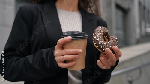 Young Caucasian with brown hair wearing black jacket drinking coffee and eating chocolate donut between skyscrapers . Outdoor, snacks time.