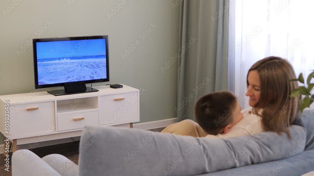 Family mother and son bonding in living room watching TV. Family time captured as they share laughter and cozy moments together. Cherish warmth of family connection in this heartwarming scene. Family mother and son bonding in living room watching TV. Family time captured as they share laughter and cozy moments together. Cherish warmth of family connection in this heartwarming scene.
