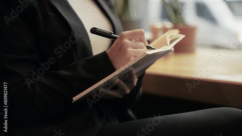 Caucasian woman in black jacket and white blouse sitting in the restaurant, drawing in her notebook, smelling and looking in the big window outside.