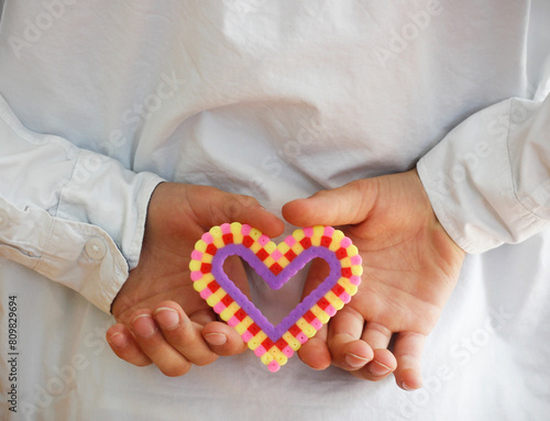 Caucasian boy's hands holding a colorful plastic heart made of ironing beads behind his back. Mother's Day and Father's Day concept with copy space.