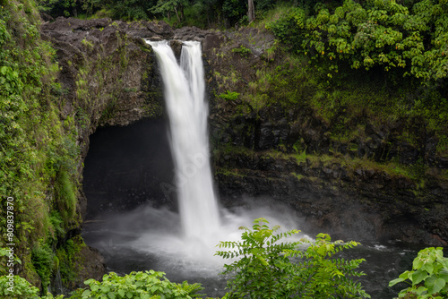 Waterfall in the rainforest, the soft, long speed of the water flowing down to the river on a mountain cave background, surrounded by green grass and trees. RAINBOW FALLS Big Island Hawaii USA