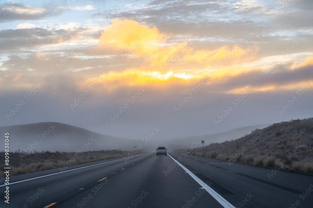 road in the mountains with raining and misty above the mountain contrast with red clouds shining by the sunset behind background, unrecognized car in the middle, Big Island Hawaii USA