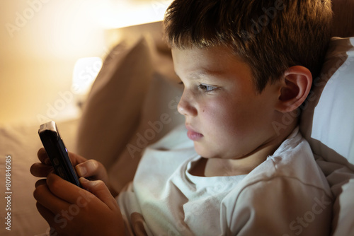 Cute little boy sitting in the dark and watching cartoons on mobile. Close up of adorable kid laying down on the bed by holding a smartphone at night. Technology concept. Copy space.