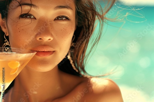 A close-up of an Asian woman with bronzed skin, slim yet curvaceous, sipping a cocktail in a bikini © Maelgoa