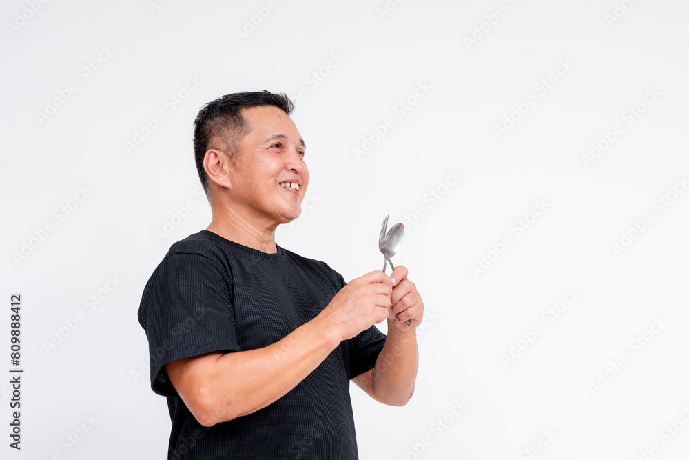 Cheerful middle aged Asian man eagerly holding a spoon and fork, dressed casually, isolated on a white background, portraying enjoyment and anticipation. Cheerful middle aged Asian man eagerly holding a spoon and fork, dressed casually, isolated on a white background, portraying enjoyment and anticipation.
