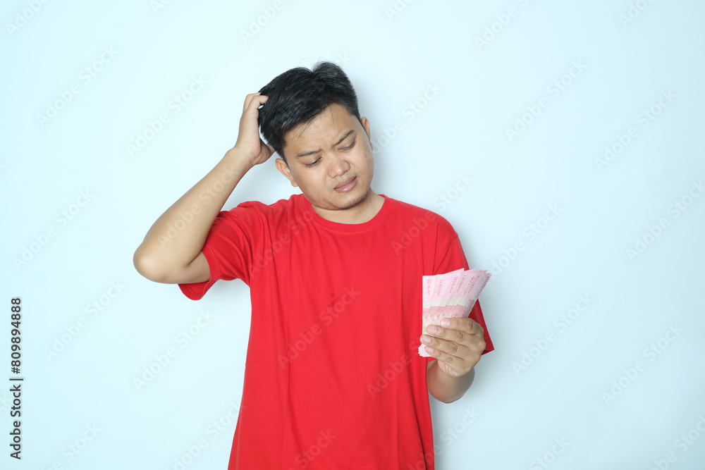 Asian man showing holding paper money and showing confused face expression. Wearing red t-shirt Asian man showing holding paper money and showing confused face expression. Wearing red t-shirt