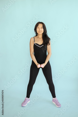 Beautiful young Asian woman doing exercises with dumbbells at biceps. Photo athletic woman with perfect body isolated on blue background. Strength and motivation.