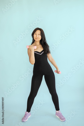 Beautiful young Asian woman doing exercises with dumbbells at biceps. Photo athletic woman with perfect body isolated on blue background. Strength and motivation.