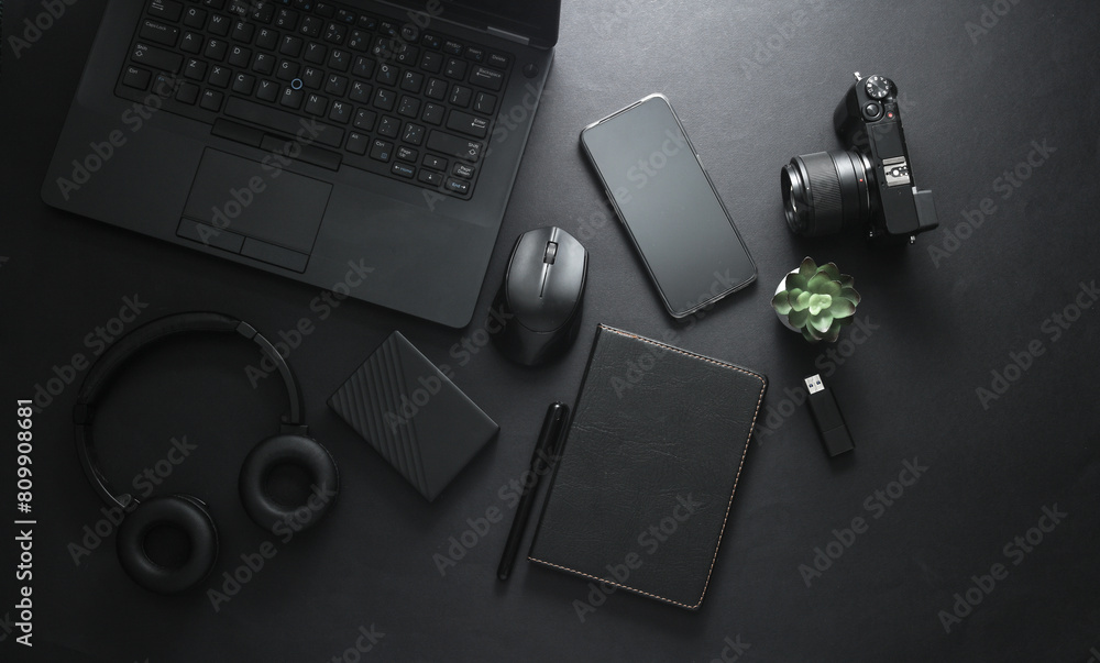 Naklejka premium Modern gadgets and accessories in black. Laptop, camera, smartphone, stereo headphones, notepad, external hard drive, USB flash drive on a black background. Working space. Flat lay. Top view