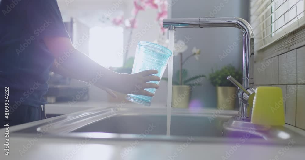 A young boy smiles as he fills a clear glass with water from a modern kitchen sink, with sunlight streaming through the window. A young boy smiles as he fills a clear glass with water from a modern kitchen sink, with sunlight streaming through the window.