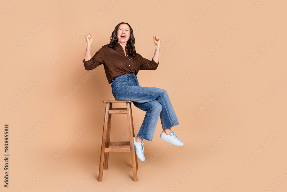 Full size photo of attractive senior woman sit chair winning dressed stylish brown clothes isolated on beige color background Full size photo of attractive senior woman sit chair winning dressed stylish brown clothes isolated on beige color background