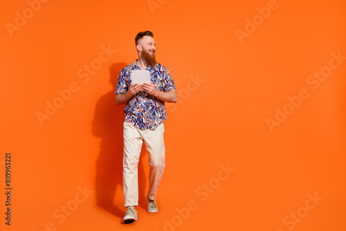 Full length photo of cool guy with red beard wear print shirt hold tablet look at offer empty space isolated on orange color background