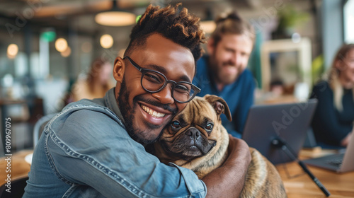 Happy black man cuddling French bulldog in the office. Candid african american male hugging office dog. Bring your pet to work day. Positive workplace culture photo