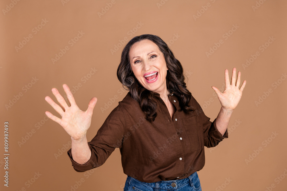 Photo portrait of attractive senior woman raise hands shocked dressed stylish brown clothes isolated on beige color background