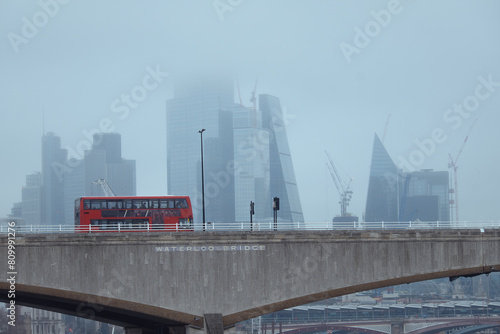 The red bus on the bridge in the foreground and the skyscrapers covered in fog. Waterloo Bridge, London, United Kingdom