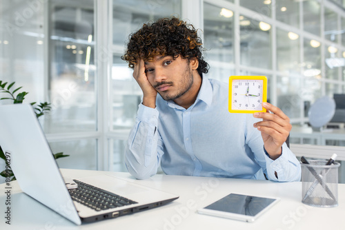 A young man sits at his office desk, displaying signs of boredom and lack of motivation as he holds a clock, signaling the slow passage of time.