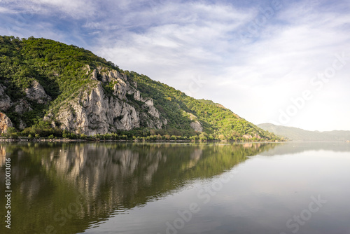the serene beauty of a natural landscape featuring a cliffside mountain with lush vegetation, reflected in the calm waters of a lake. The sky is partly cloudy