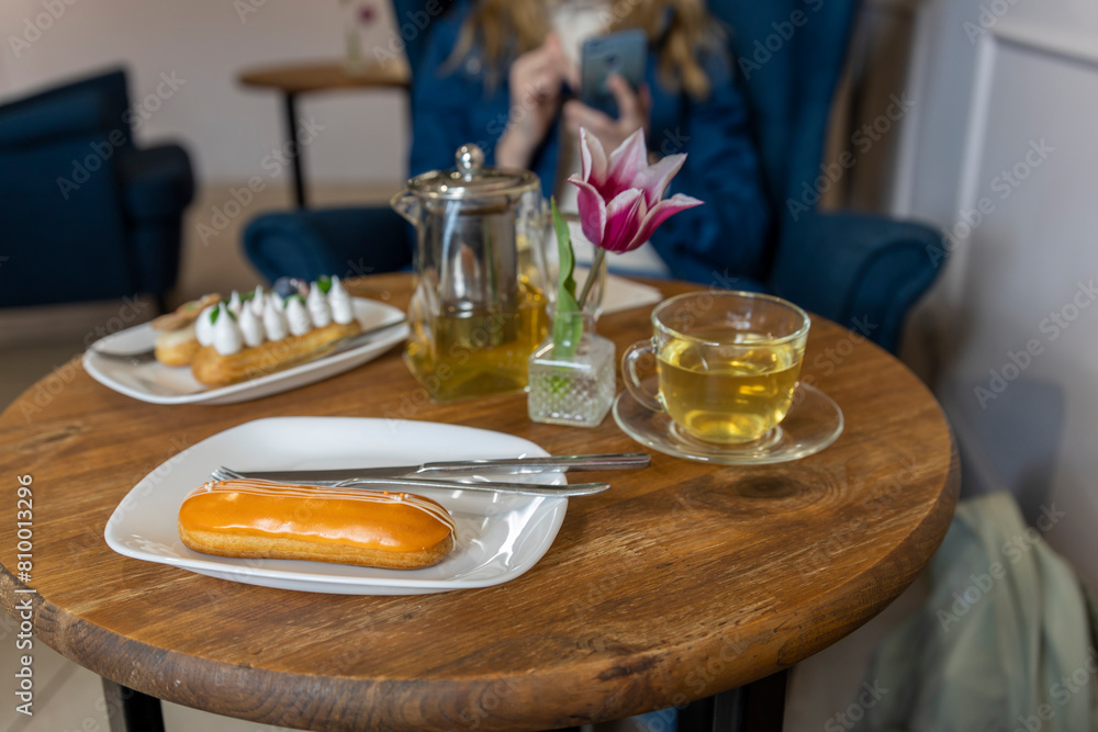 A casual café setting with a wooden table featuring an eclair on a white plate, accompanied by silver cutlery and a glass cup. A teapot and a purple tulip in a glass vase add to the ambiance A casual café setting with a wooden table featuring an eclair on a white plate, accompanied by silver cutlery and a glass cup. A teapot and a purple tulip in a glass vase add to the ambiance