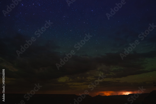 Serene night sky with numerous bright stars over a canyon or desert landscape. Dramatic contrast with clouds and distant glow at the Grand Canyon.