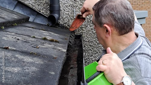 Man with green bucket on ladder cleans dirt and weeds from gutter on tiled roof