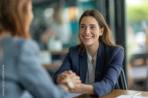 Two business women are sitting at a table, one of them smiling. They are dressed in business attire and appear to be having a conversation