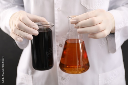 Woman holding beaker and flask with different types of crude oil on dark background, closeup