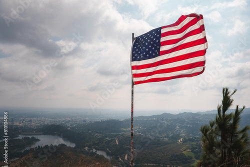 American flag waving in the wind against a hilly landscape. Flagpole on a rocky outcrop. Overcast sky with cityscape in the background, possibly Los Angeles. Water below.