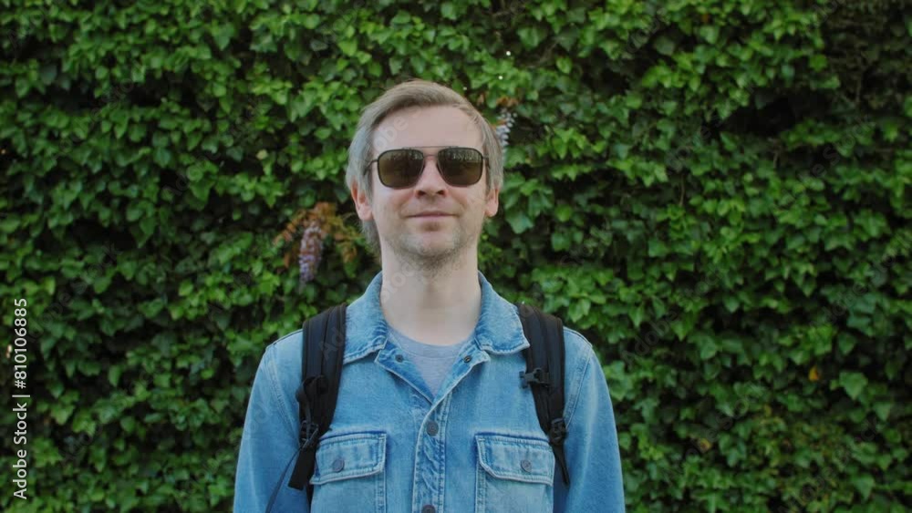 Man in sunglasses standing in front of green hedge, looking cool and leisurely Man in sunglasses standing in front of green hedge, looking cool and leisurely