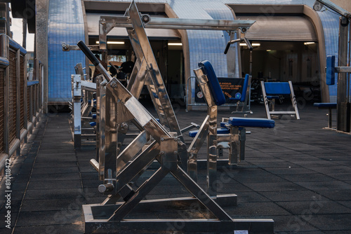 Outdoor gym area showcasing shiny metal leg press machine, blue bench, and rubber tile flooring in urban setting with unique blue tiled roof architecture. Possibly Venice Beach, Los Angeles.