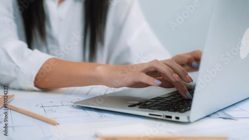 Close-up of young beautiful professional architect hands using laptop with blueprint and architectural document placed on table at modern office. Creative design concept. Focus on hand. Immaculate.