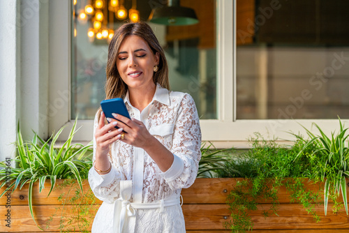Elegant and attractive woman dressed fashionably in a white suit and high heels working with the smartphone and tablet on the terrace of a cafe on the street