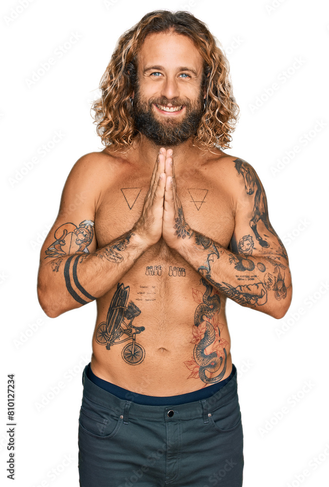 Handsome man with beard and long hair standing shirtless showing tattoos praying with hands together asking for forgiveness smiling confident. Handsome man with beard and long hair standing shirtless showing tattoos praying with hands together asking for forgiveness smiling confident.