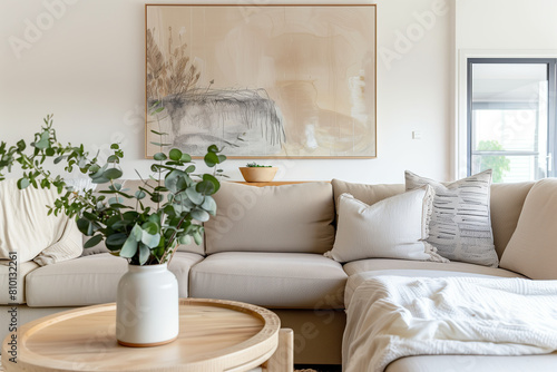 A modern living room with neutral tones, featuring an abstract painting on the wall above a beige sofa and round wooden coffee table. A vase of eucalyptus is placed next to it, adding fresh greenery.