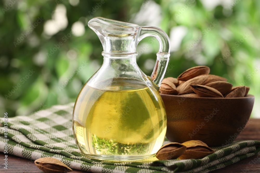 Almond oil in jug and nuts on wooden table against blurred green background, closeup Almond oil in jug and nuts on wooden table against blurred green background, closeup