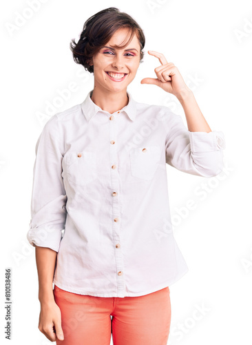 Beautiful young woman with short hair wearing elegant white shirt smiling and confident gesturing with hand doing small size sign with fingers looking and the camera. measure concept.