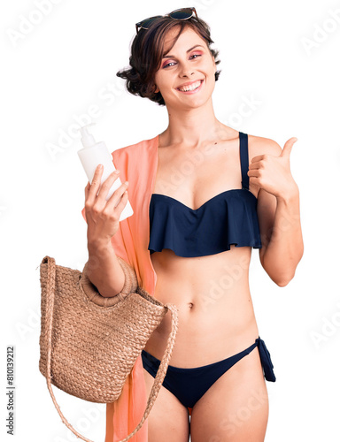 Beautiful young woman with short hair wearing bikini holding sunscreen lotion smiling happy and positive, thumb up doing excellent and approval sign