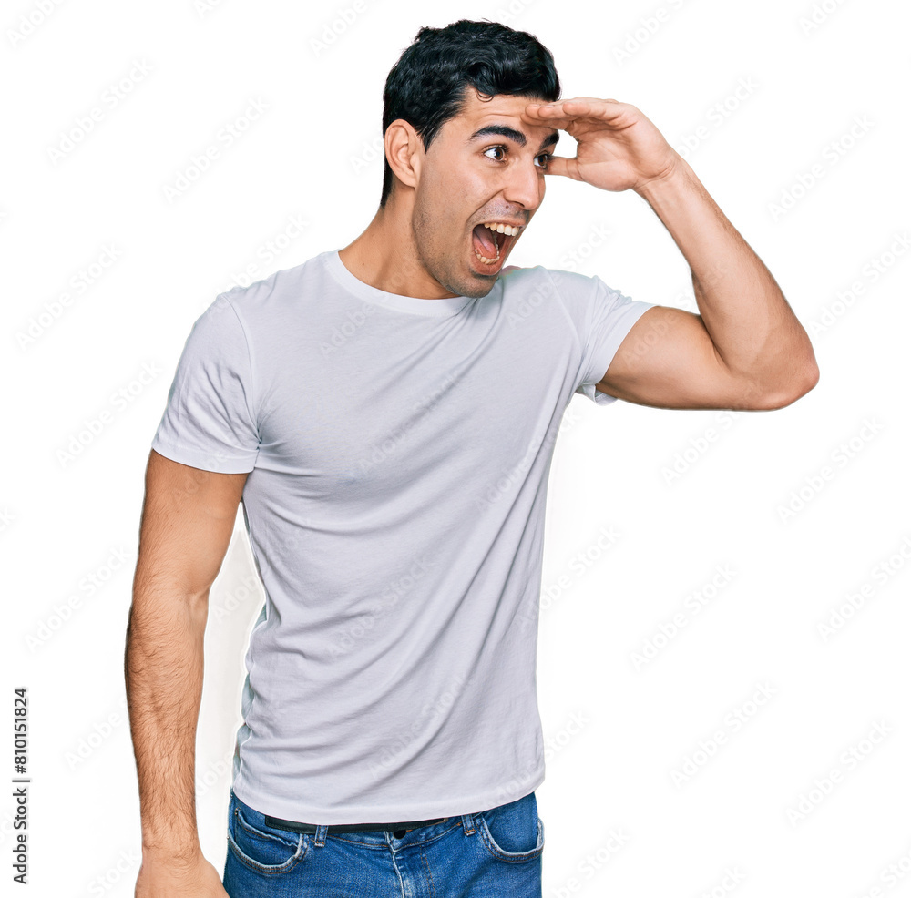 Handsome hispanic man wearing casual white t shirt very happy and smiling looking far away with hand over head. searching concept. Handsome hispanic man wearing casual white t shirt very happy and smiling looking far away with hand over head. searching concept.