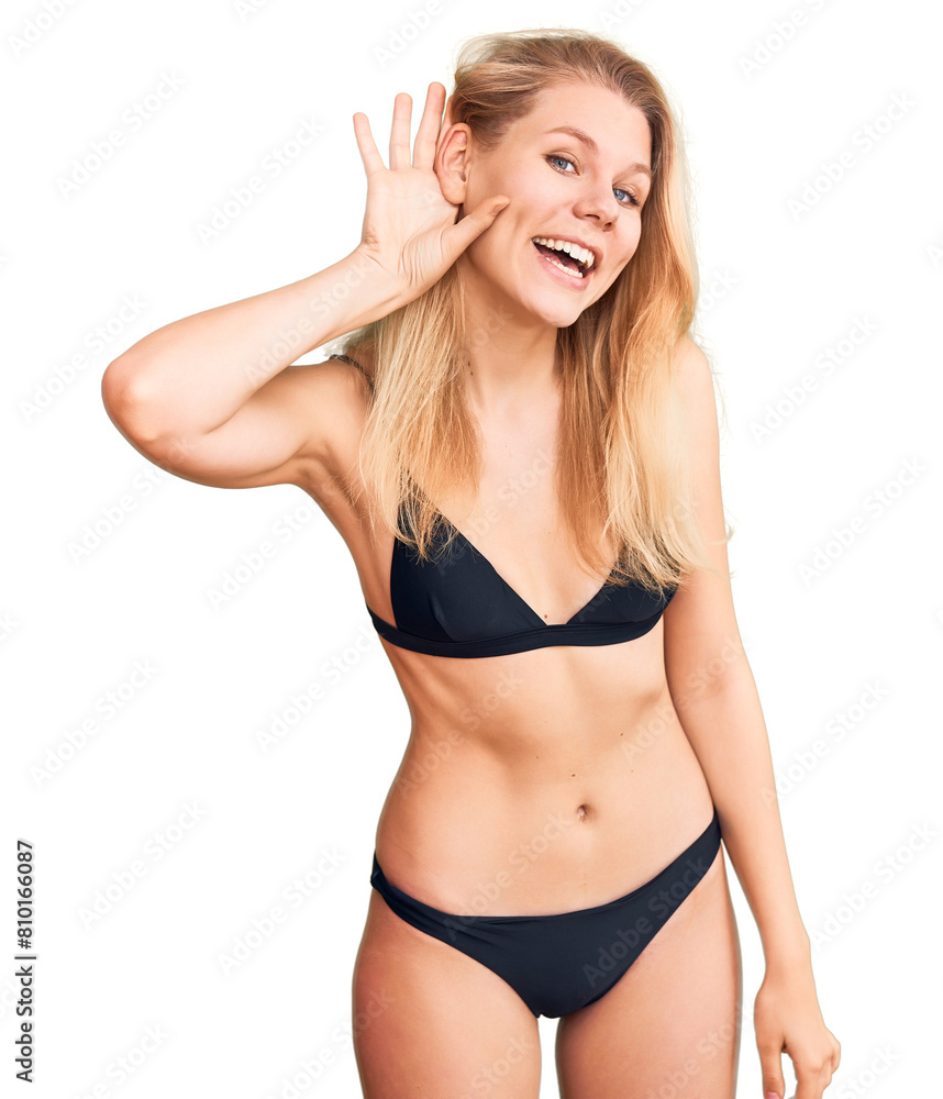 Fototapeta premium Young beautiful blonde woman wearing bikini smiling with hand over ear listening an hearing to rumor or gossip. deafness concept.