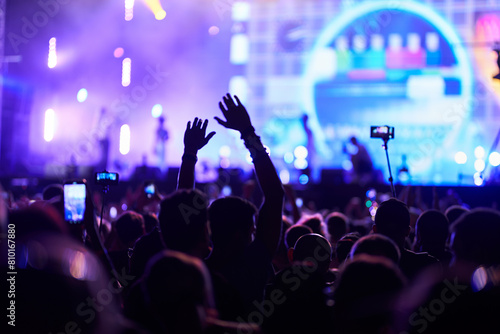 Back view of excited audience at night concert with hands raised, enjoying live music. Bright stage lights illuminate energetic fans at outdoor festival. Person records event with smartphone.